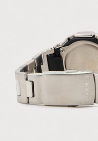 Zilverkleurige armband van roestvrij staal G-SHOCK met een matte afwerking, voorzien van een zwarte kast en een veilige sluiting. Zichtbare merkgravure op de sluiting.