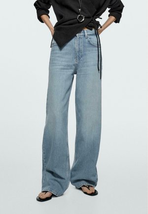 Persona con jeans azzurri a gamba larga, top nero incrociato con coulisse, collana con ciondolo e sandali infradito neri, in piedi con le mani sui fianchi.