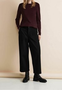 Woman wearing a dark maroon long-sleeve top, black wide-leg cropped pants with large front pockets, and black ankle boots standing against a beige wall.