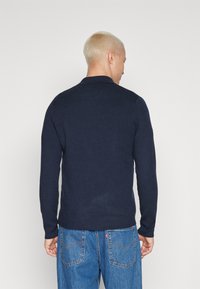 Polo de culoare bleu marin cu mânecă lungă, dintr-un material texturat, cu guler standard, purtat cu blugi deschiși la culoare, dotati cu buzunare la spate.