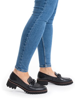 Person wearing blue skinny jeans and black chunky-heeled loafers with decorative chain detail, standing on a white background.
