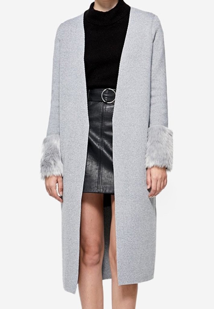 Femme portant un long manteau gris clair avec des poignets duveteux, un pull noir à col roulé et une jupe en cuir noire avec une ceinture à boucle circulaire.