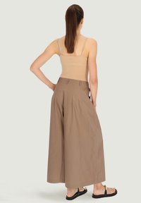 Brązowe spodnie o szerokich nogawkach, wykonane z lekkiego materiału, z plisami, wysokim stanem i gładkim wykończeniem, zestawione z płaskimi sandałami.