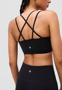 Vrouw die een zwarte sportbeha met kruisbandjes en een legging met hoge taille draagt, gezien vanaf de achterkant tegen een effen lichte achtergrond.