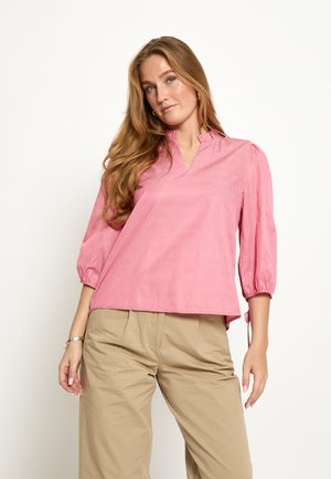 Junge Frau mit langen Haaren, die eine rosa Bluse mit dreiviertel Ärmeln und beige Hosen trägt, vor einem schlichten weißen Hintergrund stehend.