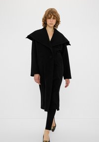 Cappotto di lana nero con un ampio colletto a scialle, chiusura a un bottone, vestibilità ampia, tasche laterali, abbinato a leggings e scarpe nere.