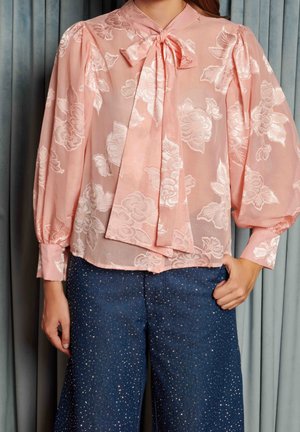Vrouw draagt een doorschijnende roze bloemige blouse met een strik en pofmouwen, gecombineerd met donkerblauwe glinsterende broek.