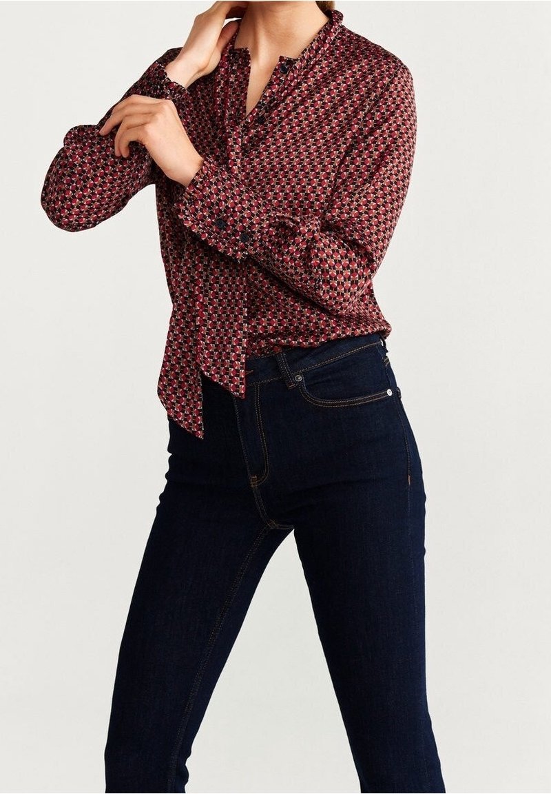 Rød og marineblå mønstret bluse med krave og bindedetalje, lavet af letvægtigt stof, bæres med mørke denimjeans. Lange ærmer er samlet ved manchetterne.