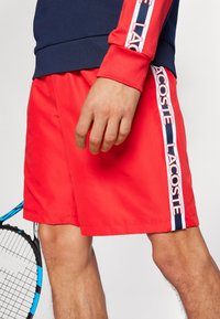 Sarkanas sporta bikses ar gludu tekstūru, sānu svītrām ar "LACOSTE" zīmolu zilā un baltā krāsā, uzvilktas ar tumši zilas krāsas topu un tenisa raketi.