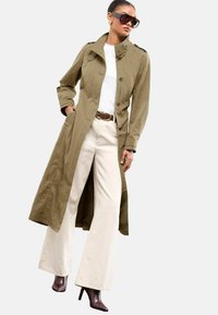 Vrouw in een olijfkleurige trenchcoat, witte t-shirt, crèmekleurige wijde broek, bruine hoge laarzen, zonnebril en gouden oorbellen.