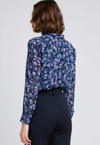 Blouse noire à fleurs avec des fleurs bleues et blanches, manches longues et col volanté, accompagnée de pantalons foncés taille haute.