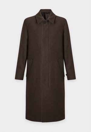 Un homme se tient debout, portant un long manteau marron à trois boutons, un pantalon foncé, des chaussures marron habillées et une chemise blanche, les mains dans les poches du manteau.