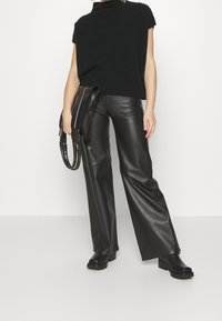 Pulsera corta negra y pantalones de cuero negros de cintura alta, que destacan piernas anchas y una textura suave. Calzado negro voluminoso completa el look.