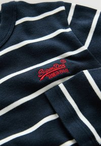 Tummansininen neulepaita, jossa on valkoisia vaakaraitoja, ribbauskaulus ja punainen brodeerattu "SuperDry" -logo edessä.