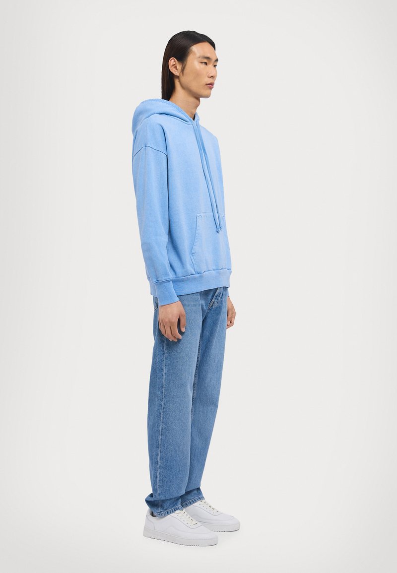 Felpa leggera azzurra con cappuccio regolabile, tasca frontale e polsini a coste, abbinata a jeans azzurri larghi e sneakers bianche.