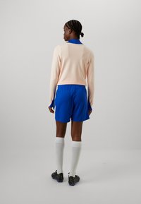 Šviesiai rožinė ilgomis rankovėmis marškinėliai su mėlynomis detalėmis, mėlynos šortai, balti iki kelio kojinės ir juodi futbolo bateliai. Lygus audinys su sportiniu pritaikymu.