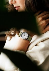 Naine toetab pead käele, kandes rohelise rihmaga käekella, millel on valge kellanumber ja mustad numbrid, osaliselt varjatud tumedate lehtede taha.