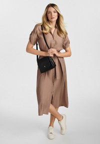 Brązowa sukienka o długości do kolan, z krótkimi rękawami i guzikami z przodu. Z akcentem w postaci związanej talii. Połączona z białymi trampkami i czarną torbą.