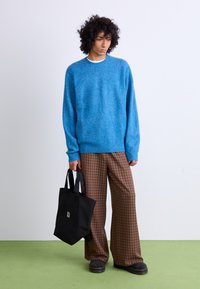 Pull en maille bleu texturé, pantalon large à carreaux marron, sac fourre-tout noir et chaussures noires épaisses, sur un fond uni.