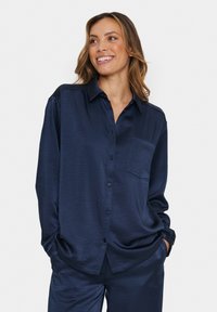 Navy satin button-up shirt with a collar, single chest pocket, and loose fit. Features subtle texture and long sleeves with cuff detail.