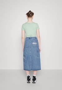 Osoba s copy ve vlasech, která má na sobě mátově zelené tričko, džínovou sukni střední délky s nášivkami a černé vysoké tenisky, stojící zády.