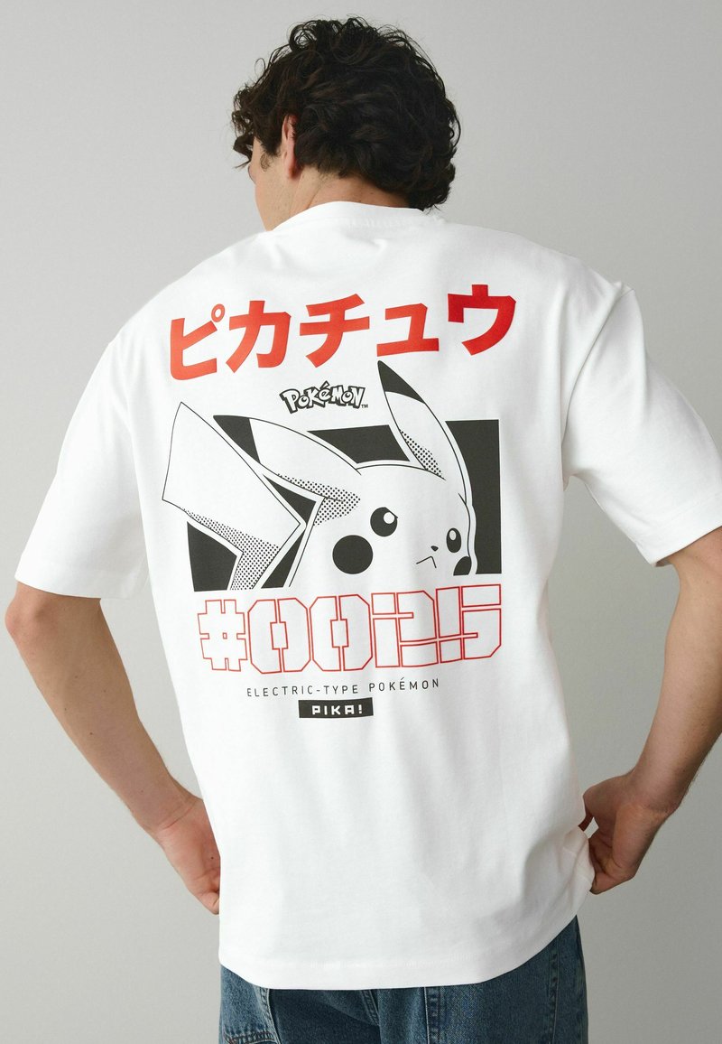 Homme portant un t-shirt blanc avec un grand graphique noir et blanc de Pikachu et un texte japonais rouge au dos, assorti à un jean bleu.