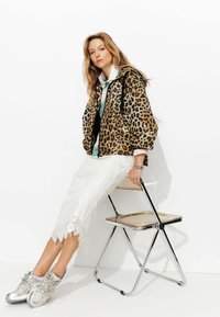 Veste oversize imprimé léopard, jupe en dentelle blanche avec un ourlet à franges, et baskets argentées métalliques. Assise sur une chaise pliante en chrome.