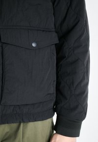 Zwarte gewatteerde jas met een drukknoopflapzak, ribgebreide manchetten en een matte afwerking. De jas heeft een textiele stof en een gestroomlijnd ontwerp.