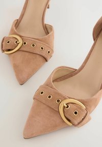 Scarpe con tacco alto in camoscio beige, con punte appuntite, dotate di una cinturini decorativo con hardware dorato e dettagli con occhielli.