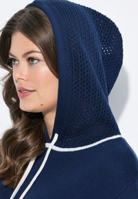 Sweat à capuche tricoté marine avec une capuche en tricot texturé ouvert, comportant un cordon blanc et un corps en tissu lisse. Vue de profil en gros plan.