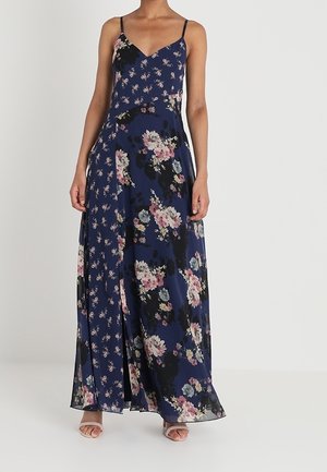 Kvinde iført en mørkeblå maxikjole med spaghetti stropper og pink og hvide blomsterprint, stående mod en ensfarvet baggrund.