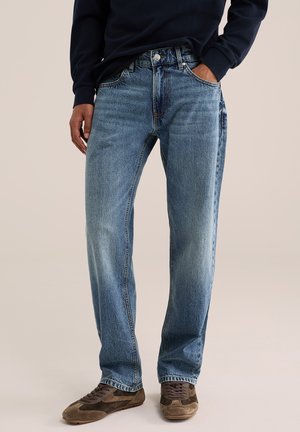 Personne portant un jean droit bleu, un sweat-shirt foncé et des baskets marron, une main dans la poche, sur un fond neutre.