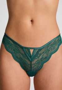 Frau trägt grüne Spitzenunterwäsche im Bikini-Stil, die ein detailliertes florales Spitzenmuster und ein Ausschnittdesign vorne zeigt.