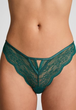Femme portant des sous-vêtements en dentelle verte de style bikini, montrant un motif floral détaillé et un design découpé à l'avant.