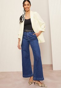 Svetle žlutý blazer přes černý top, doplněný širokými modrými džíny. Akcentováno vzorovanými podpatky. Jednoduchý design a ležérní vzhled.