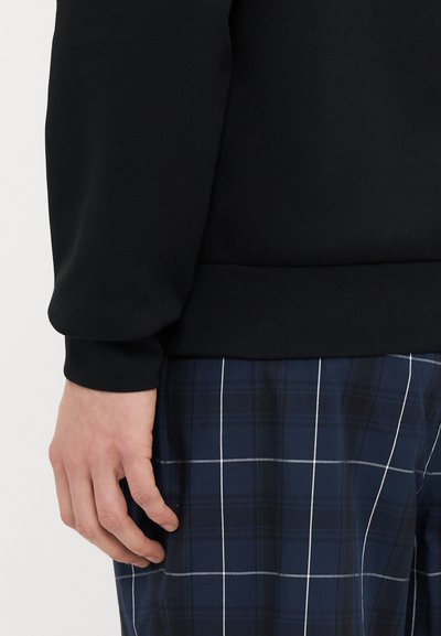 Sweatshirt noir en tissu texturé avec une taille ajustée, associé à un pantalon à carreaux bleu marine avec des lignes blanches et une coupe décontractée.