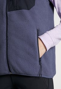 Person, die eine lila Fleece-Weste mit einer schwarzen Reißverschlusstasche trägt, mit der Hand in der rechten Tasche, über einem helllila Langarmoberteil.