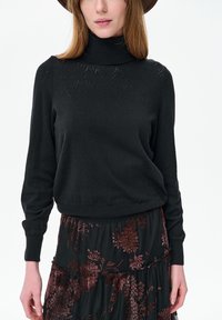 Vrouw die een zwarte coltrui met subtiele gebreide patronen draagt en een zwarte rok met donkerrode bloemmotieven, staand tegen een witte achtergrond.