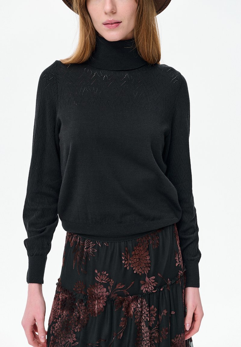 Vrouw die een zwarte coltrui met subtiele gebreide patronen draagt en een zwarte rok met donkerrode bloemmotieven, staand tegen een witte achtergrond.