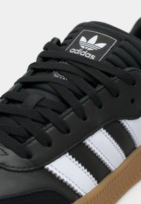 Svart skinnsneaker med tre hvite striper, teksturerte lisser, polstret krage og en gummisåle med logoetikett på tungen.