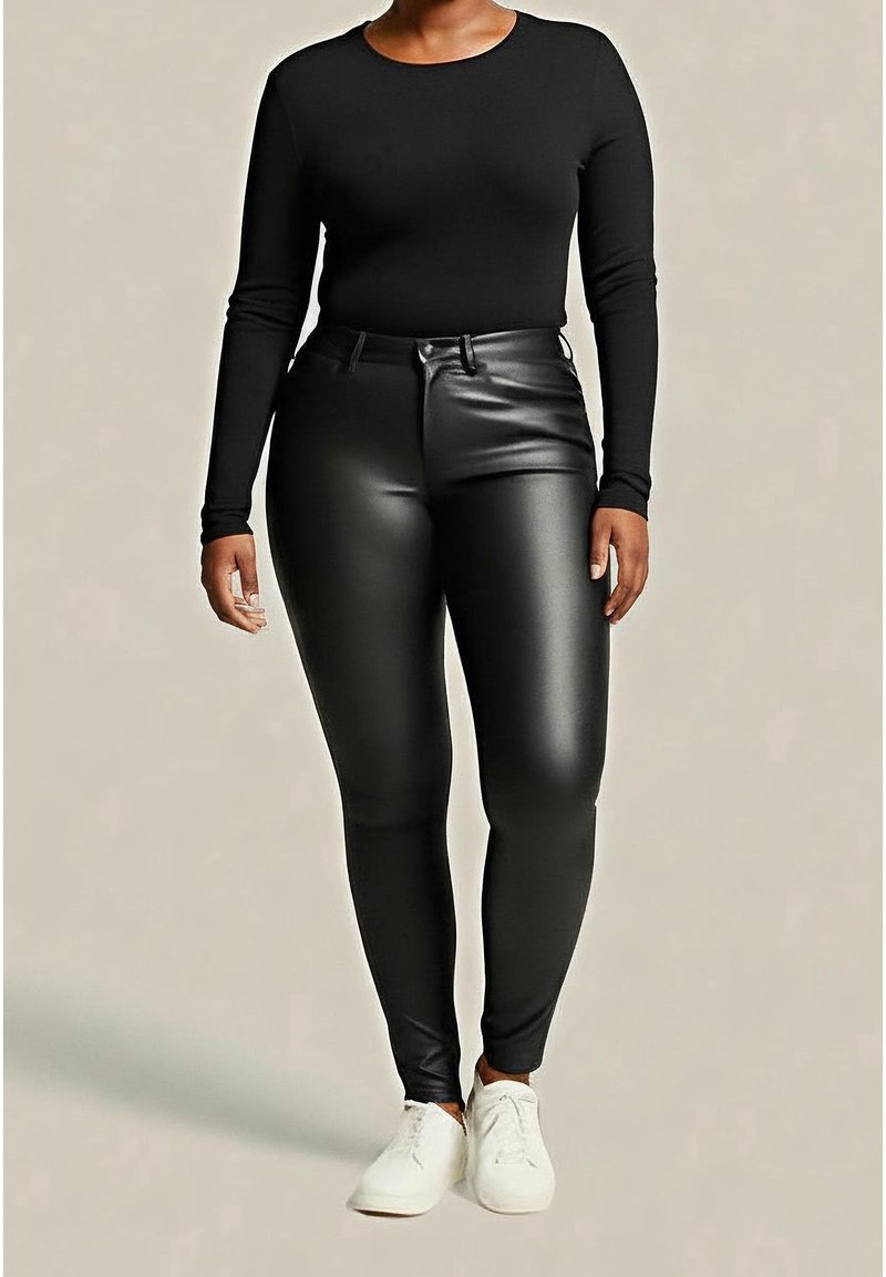Personne portant un t-shirt noir à manches longues, un pantalon noir en simili-cuir taille haute et des baskets blanches, debout devant un fond uni.