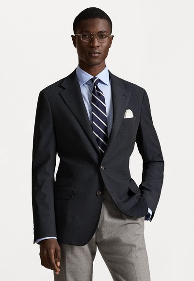 Σκούρο κομμένο σακάκι με κολάρο notch, συνδυασμένο με ανοιχτό μπλε πουκάμισο και σκούρη ριγέ γραβάτα. Γκρι παντελόνι, λευκή μαντήλα και γυαλιά.