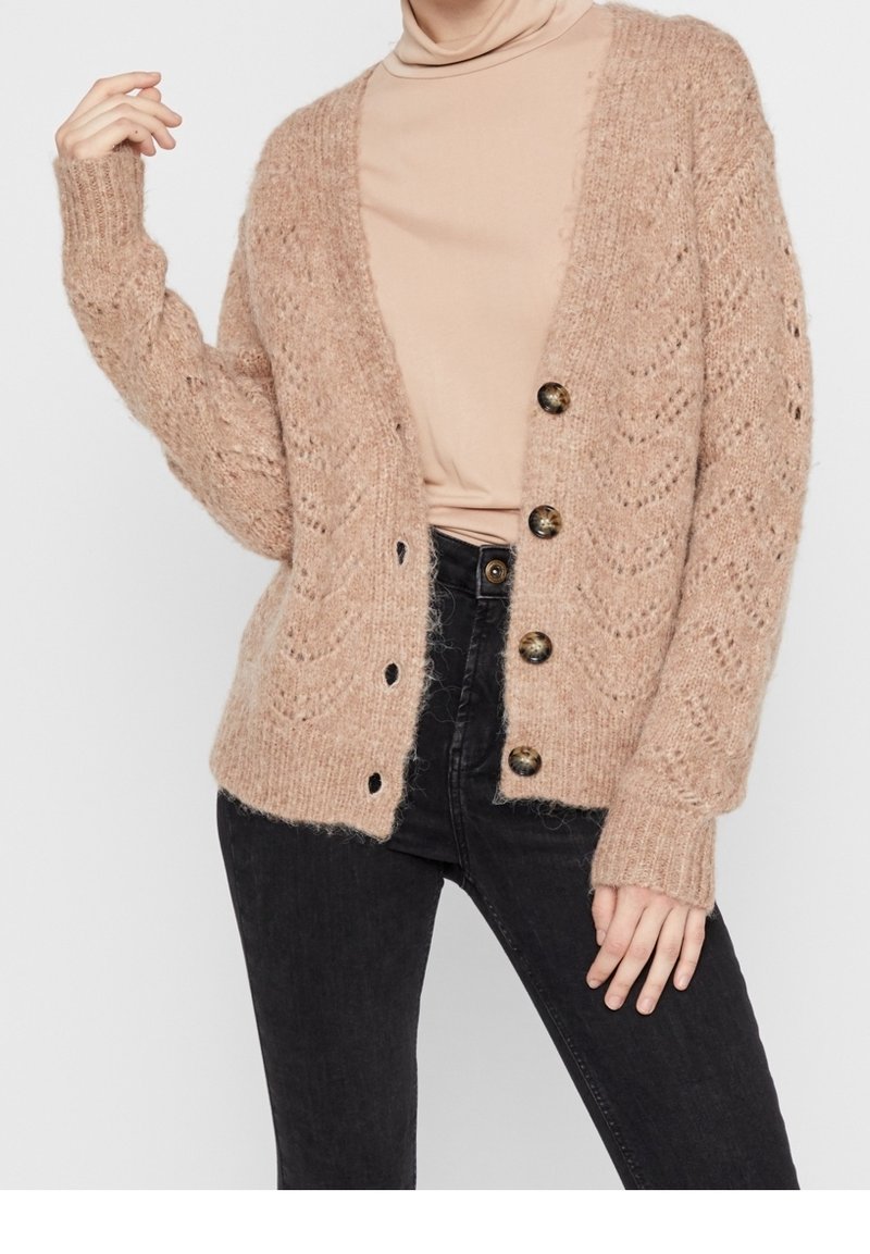 Femme portant un cardigan beige en maille avec des boutons en écaille de tortue sur un col roulé beige, assorti à un jean noir taille haute.