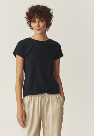 Žena s krátkymi kučeravými hnedými vlasmi, oblečená v čiernom tričku s krátkym rukávom a béžových nohaviciach so šnúrkou v páse, stojaca s jednou rukou v vrecku.