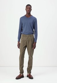 Dunkelblauer V-Ausschnitt-Pullover, grüne Chinos und braune Lederschuhe. Der Pullover ist weich; die Chinos haben einen lockeren Schnitt und eine subtile Textur.