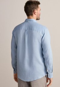 Camisa de manga larga azul claro hecha de una tela suave y texturizada. Presenta un collar de punta, puños abotonados y un dobladillo curvado con un detalle de plisado.