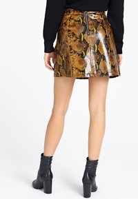 Donna che indossa una mini gonna in pelle di serpente marrone con zip sul retro e stivaletti neri con tacco, in piedi su uno sfondo bianco.