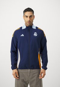 Mørkeblå treningsjakke med lyseblå detaljer, hvite sidelapper og oransje striper. Har full glidelås og brodert logo.
