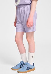 Barn iført lyse lilla sportsshorts, hvide crew-sokker med logo og blå sneakers med gummisåler, står mod en hvid baggrund.