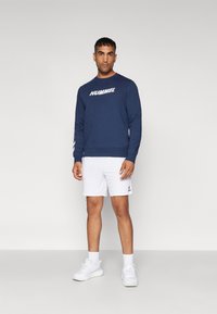 Mørkeblå sweatshirt med hvid "HUMMEL" tekst; parret med hvide shorts og hvide sneakers. Enkelt farve, rund hals, lange ærmer.